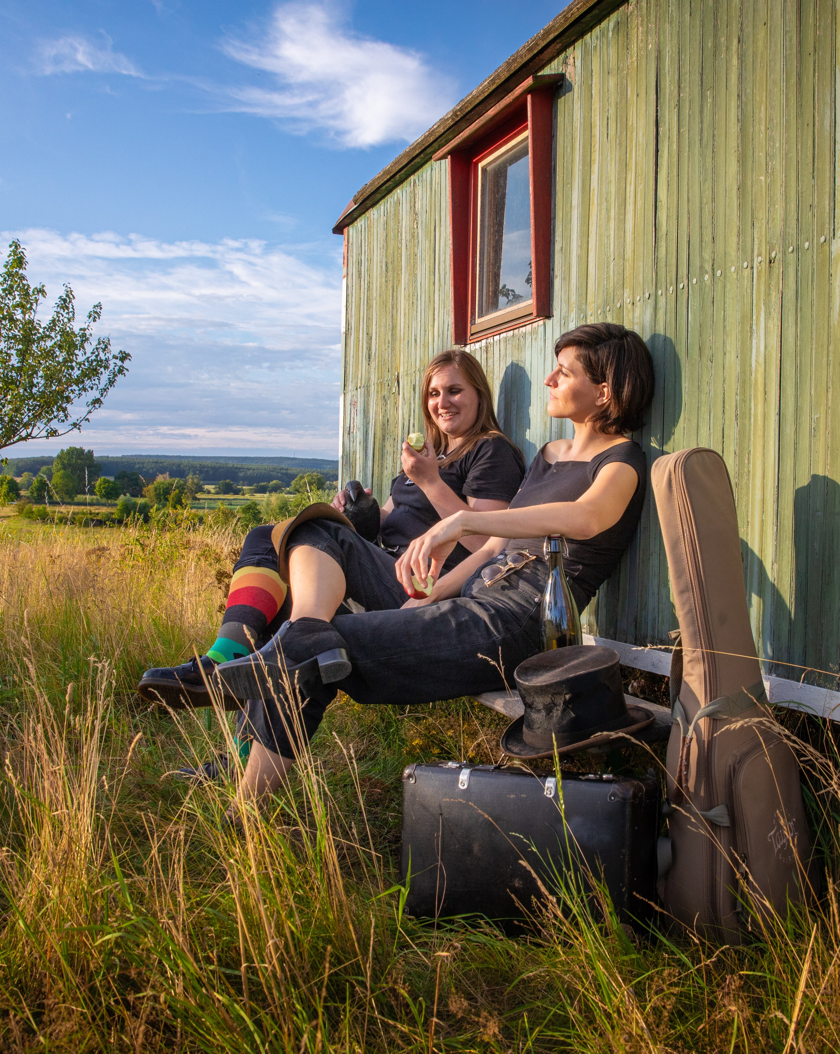 Bild vergrößern: 2 Frauen sitzen vor einem alten Wohnwagen auf zwei Klappstühlen. Mitten in einem Feld. Vorne ist eine Gitarre in einem Gitarrencase zu sehen, ein Zylinder-Hut auf einem Koffer. Eine Der Frauen isst einen Apfel.