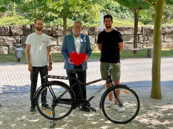 Drei Personen stehen hinter einem Fahrrad. Die Person in der Mitte hat ein rotes Plschherz in der Hand.