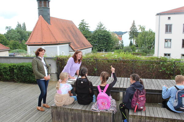 Bild vergrößern: Die Kinder sitzen auf Holzbänken vor dem Holztisch und die beiden Gästeführerinnen haben ein Lächeln im Gesicht.