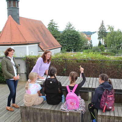 Bild vergrößern: Die Kinder sitzen auf Holzbnken vor dem Holztisch und die beiden Gstefhrerinnen haben ein Lcheln im Gesicht.