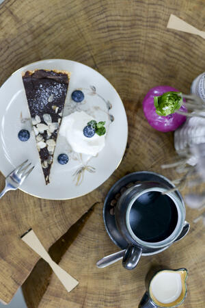 Bild vergrößern: Auf den weißen Teller befindet sich ein Schokoladenkuchen mit Schlagsahne, garniert mit Heidelbeeren. Daneben steht eine Tasse schwarzer Kaffee.
