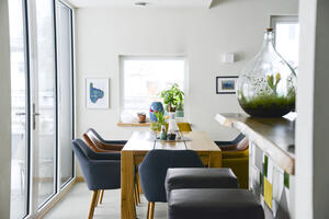 Bild vergrößern: An dem großen Holztisch gibt es blaue und braune Ledersessel. Am rechten Seitenrand ist noch eine Bar mit zwei schwarzen Hockern zu erkennen. Dekoriert ist der Tisch mit einer weißen Blumenvase.