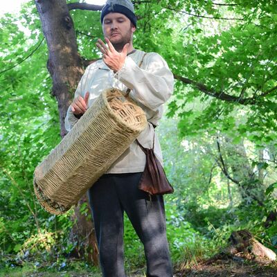 Bild vergrößern: Der Fischer steht unter einen grnen Baum, mit seiner Reuse, und hebt seine linke Hand erklrend an.