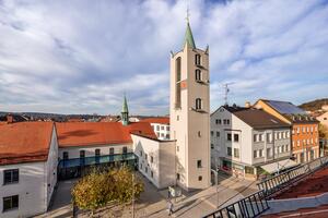 Bild vergrößern: Unter den blauen wolkenverhangenen Himmel streckt sich weit nach oben der Turm der Erlserkirche. Neben der Kirche finden sich weitere Huser an der Bahnhofsstrae entlang. Auf dem Vorplatz der Kirche stehen Bume und Bnke.