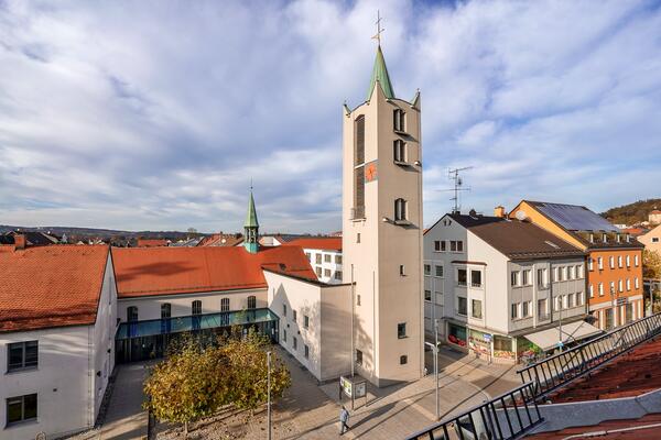 Unter den blauen wolkenverhangenen Himmel streckt sich weit nach oben der Turm der Erlserkirche. Neben der Kirche finden sich weitere Huser an der Bahnhofsstrae entlang. Auf dem Vorplatz der Kirche stehen Bume und Bnke. 