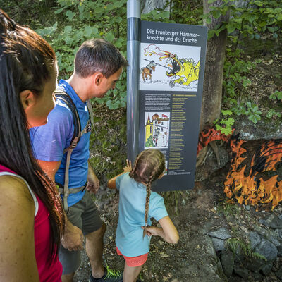 Das kleine M&auml;dchen im blauen Shirt steht vor der gro&szlig;en Tafel und liest ihren Eltern die Sage von der Drachenh&ouml;hle vor. Im Hintergrund erkennt man den Drachen aus Blech, der in schwarz und gelb angemalt ist.
