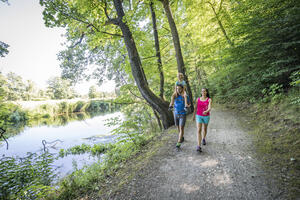 Bild vergrößern: Der Vater trägt seine Tochter auf den Schultern und die Mutter geht nebenher. Die Drei sind bekleidet mit Shorts und Shirts. Der Weg ist entlang der Naab schattig. Auffällig ist der alte große Baum im Hintergrund.