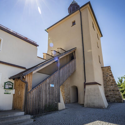Unter blauen Himmel steht mächtig der Blasturm mit seiner überdachten Holztreppe. Er wird durch feste Sandsteinmauern gestützt.