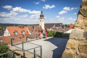 Bild vergrößern: Im Hintergrund erhebt sich der Kirchturm unter blauen Himmel. Auf der Plattform steht eine Familie und blickt über die roten Dächer der Stadt.