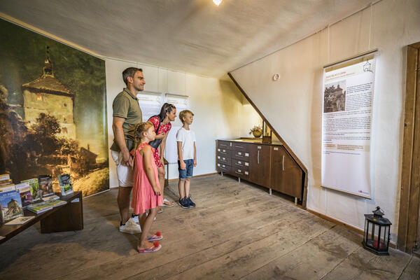 Die Eltern stehen mit dem Sohn und der Tochter im Raum und lesen das Banner an der Wand. Links daneben befindet sich eine Vitrine aus Holz. Hinter der Familie hängt ein Bild an der Wand.