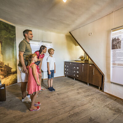 Die Eltern stehen mit dem Sohn und der Tochter im Raum und lesen das Banner an der Wand. Links daneben befindet sich eine Vitrine aus Holz. Hinter der Familie hängt ein Bild an der Wand.