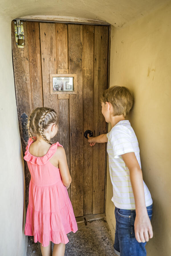 Bild vergrößern: Vor der Holztür stehen zwei Kinder, ein Mädchen und ein Bub, und horchen der Aufnahme des Knödelliedes zu.