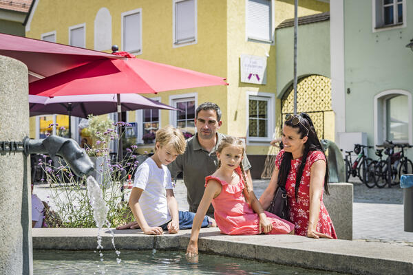 Im Hintergrund befindet sich ein gelbes Haus und rote Sonnenschirme spenden Schatten. Die kleine Familie erfrischt sich am Brunnen.



