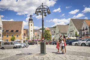 Bild vergrößern: Im Vordergrund steht ein Glockenspiel.  Interessiert schaut eine Familie zu den zehn Glocken nach oben. Im Hintergrund steht prächtig die Kirche unter einem blauen Himmel, durchzogen mit ein paar weißen Wolken.