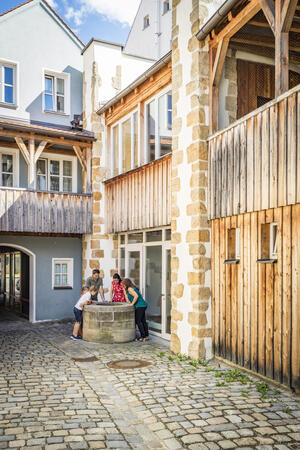 Bild vergrößern: Auf der rechten Seite befindet sich ein Haus, welches noch zum Teil mit Holz verkleidet ist. In der Ecke befindet sich ein Brunnen und die kleine Gruppe seht rundherum und blickt hinein. Ein schöner Holzbalkon ziert das blaue Haus im ersten Stock.