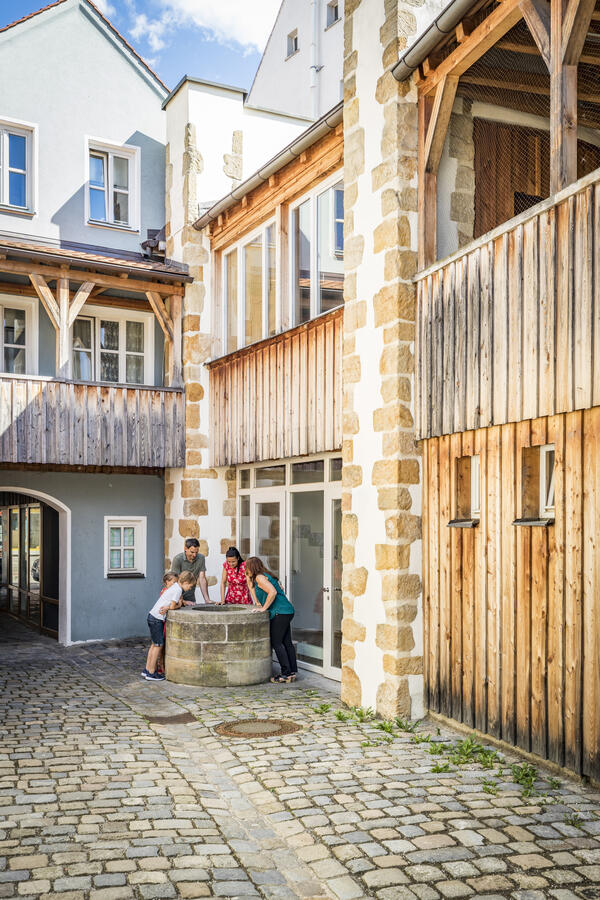 Bild vergrößern: Auf der rechten Seite befindet sich ein Haus, welches noch zum Teil mit Holz verkleidet ist. In der Ecke befindet sich ein Brunnen und die kleine Gruppe seht rundherum und blickt hinein. Ein schöner Holzbalkon ziert das blaue Haus im ersten Stock.