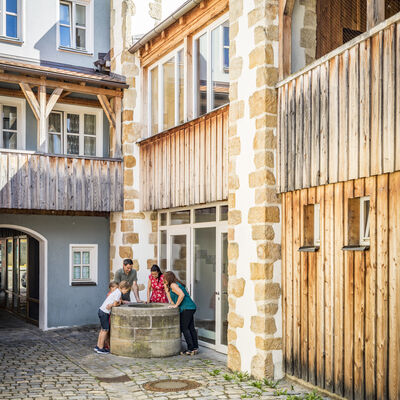 Bild vergrößern: Auf der rechten Seite befindet sich ein Haus, welches noch zum Teil mit Holz verkleidet ist. In der Ecke befindet sich ein Brunnen und die kleine Gruppe seht rundherum und blickt hinein. Ein schöner Holzbalkon ziert das blaue Haus im ersten Stock.