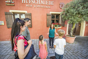 Bild vergrößern: Die Gästeführerin steht vor einem roten Haus mit dem Namen "Hufschmiede - Wein-Speiselokal" einer kleinen Besuchergruppe gegenüber.