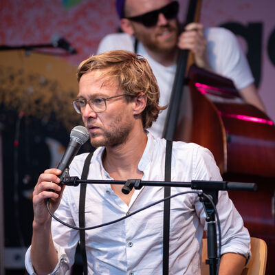 Portrait eines Künstlers im weißen Hemd mit Mikrofon auf der Bühne.