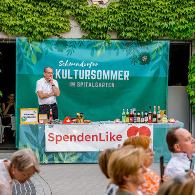 Ein Getränkestand, der mit grünen Bannern und der Aufschrift "SpendenLike" dekoriert ist. Dahinter steht ein Mann in weißem Hemd.