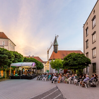 Seitlicher Blick auf eine beleuchtete Bühne mit weißem Dach und das davor sitzende Publikum. Im Hintergrund sind ein heller Abendhimmel, eine Kapelle und ein Kran abgebildet.