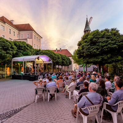 Zuschauer sitzen in einem von Bäumen umrahmten Platz im Abendlicht auf Plastikstühlen und hören einer Band zu, die auf einer bunt beleuchteten Bühne mit weißem Dach spielt.