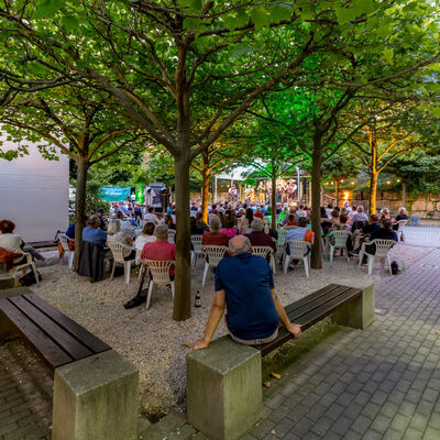 Menschen sitzen auf weißen Plastikstühlen zwischen Platanen und lauschen einem Konzert der Van Straaten Band. Im Vordergrund sitzt ein Mann in einem blauen Shirt auf einer Holzbank und hört ebenfalls zu.