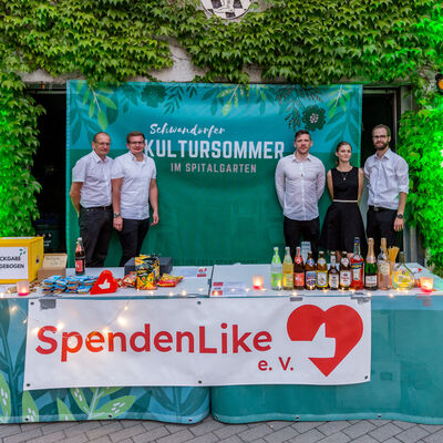 Ein Getränkestand, vor dem ein rot-weißes Banner mit der Aufschrift "SpendenLike e.V." aufgehängt ist. Hinter dem Stand stehen drei Männer in weißen Hemden und eine Frau in einem schwarzen Kleid.