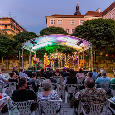 Die Kapelle So und So spielt auf einer bunt beleuchteten Bühne. Davor sitzt das Publikum.