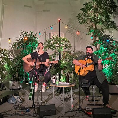 Die Musiker Susi Raith und Mathias Kellner spielen auf eine Bühne Gitarre und singen. Die Bühne ist mit vielen Pflanzen und bunten Lichtern dekoriert.