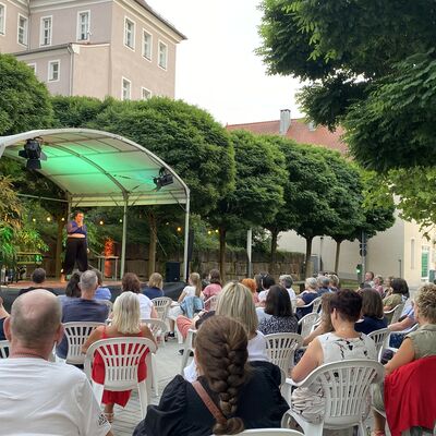 Menschen in weißen Plastikstühlen blicken zu der Kabarettistin Teresa Reichl, die auf einer bunt beleuchteten Bühne steht und in ein Mikrofon spricht.
