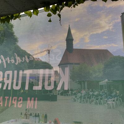 Spitalkirche, Bühne im Spitalgarten und das Publikum davor durch ein Werbetransparent hindurch bei Dämmerung fotografiert.