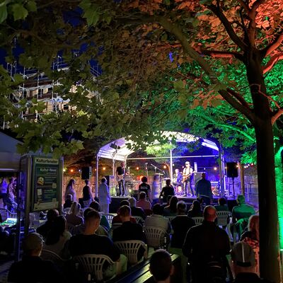 Sitzende und stehende Zuschauer blicken auf eine bunt beleuchtete Bühne, auf der eine Rockband spielt.