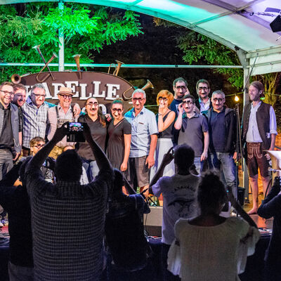 Die Band steht mit einigen Fans, die Faschingsbrillen mit falschen Nasen tragen auf der Bühne. Mehrere Menschen schließen ein Foto von ihnen.