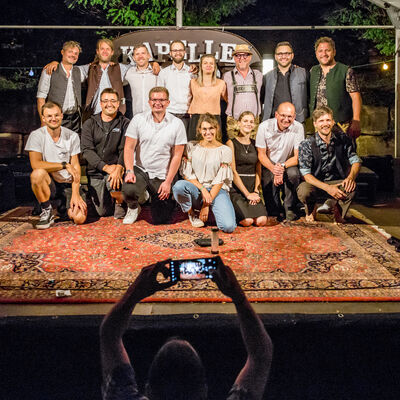 Gruppenfoto von der Band, den Veranstaltern und den Helfern auf der beleuchteten Bühne. Davor schießt ein Mann ein Foto mit dem Handy.