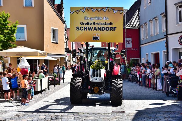 Bild vergrößern: Ein Bulldog fährt als Teil eines Festzuges die Straße entlang. Darauf ist ein großes gelbes Schild mit der Aufschrift "Es grüßt die große Kreisstadt Schwandorf" montiert.