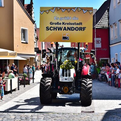 Ein Bulldog fährt als Teil eines Festzuges die Straße entlang. Darauf ist ein großes gelbes Schild mit der Aufschrift "Es grüßt die große Kreisstadt Schwandorf" montiert.