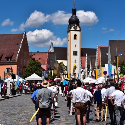 Mehrere Menschen in Tracht gehen, von hinten fotografiert, als Teil eines Festzugs auf einen weißen Kirchturm zu.