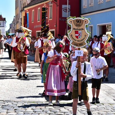 Teilnehmende eines Festzuges. Vorne eine Kindergruppe in Tracht, ein Kind ganz vorne hält eine Holztafel in die Höhe. Hinten geht eine spielende Blaskapelle.
