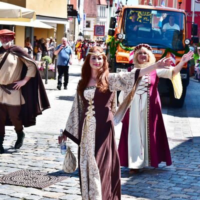 Teilnehmende eines Festzugs. Vorne gehen zwei Frauen und ein Mann in mittelalterlichen Gewändern. Hinten fährt ein Feuerwehrauto.