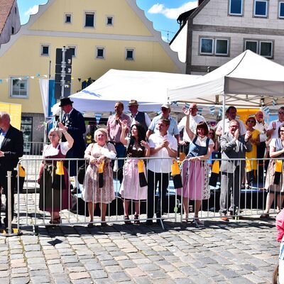 Mehrere Menschen in Tracht stehen hinter einer halbhohen Absperrung. Hinter ihnen stehen zwei weiße Pavillons.