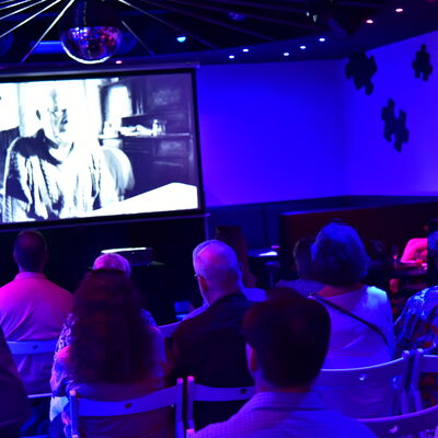 Menschen sehen einen schwarz-weiß Film auf einer großen Leinwand in einer blau ausgeleuchteten Diskothek.