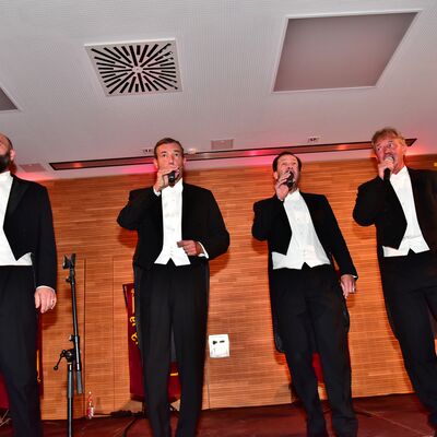Vier Männer in schwarzen Anzügen und weißen Hemden singen in Mikrophone.