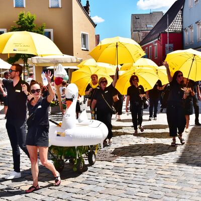 Mehrere schwarz gekleidete Menschen mit großen gelben Regenschirmen gehen als Teil eines Festzugs die Straße entlang. Sie ziehen einen großen aufblasbaren Schwan auf einem Rollwagen mit sich.