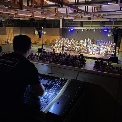 Ein Mann an einem Mischpult steuert den Ton für ein Chorkonzert in einer Turnhalle im hinteren Teil des Bildes.