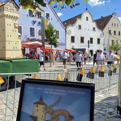Ein Bauzaun, der über einen Marktplatz verläuft, trennt einen Festzug im hinteren Teil, von dem vorderen Teil. Dort steht präsent ein Aufsteller mit einem bunten Plakat.