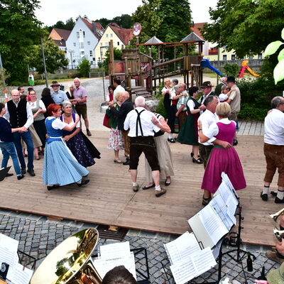 Mehrere Paare tanzen in Tracht im Freien auf einem Tanzboden. Sie werden von einer Blaskapelle begleitet.