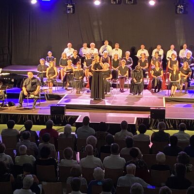 Ein großer Chor sitzt auf einer beleuchteten Bühne in einer Turnhalle. Davor sitzt das Publikum.