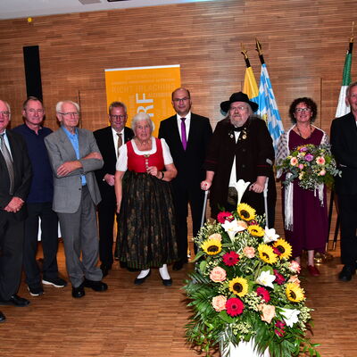 Mehrere festlich gekleidete Menschen stehen im Halbkreis um ein großes Blumengesteck. Eine Frau hält einen Blumenstrauß.