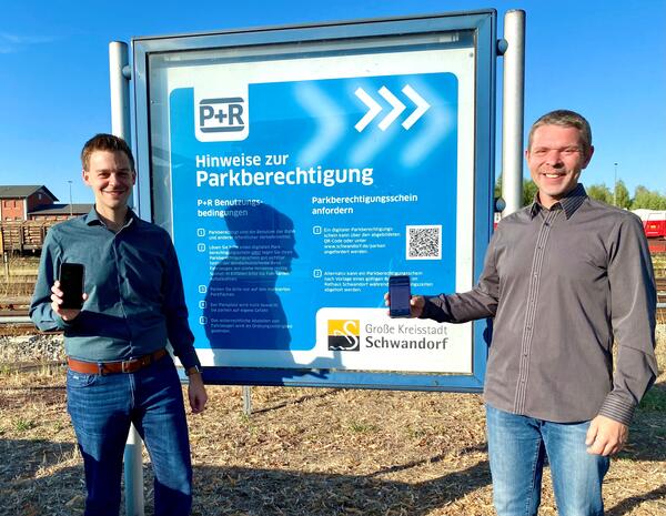 Bild vergrößern: Das Bild zeigt zwei Männer vor einer beschrifteten Tafel. Jeder der Männer hat ein Handy in der Hand.